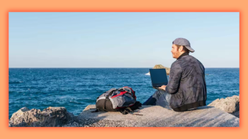 a person setting on a rock on a beach with a laptop in his lap while looking at the sea